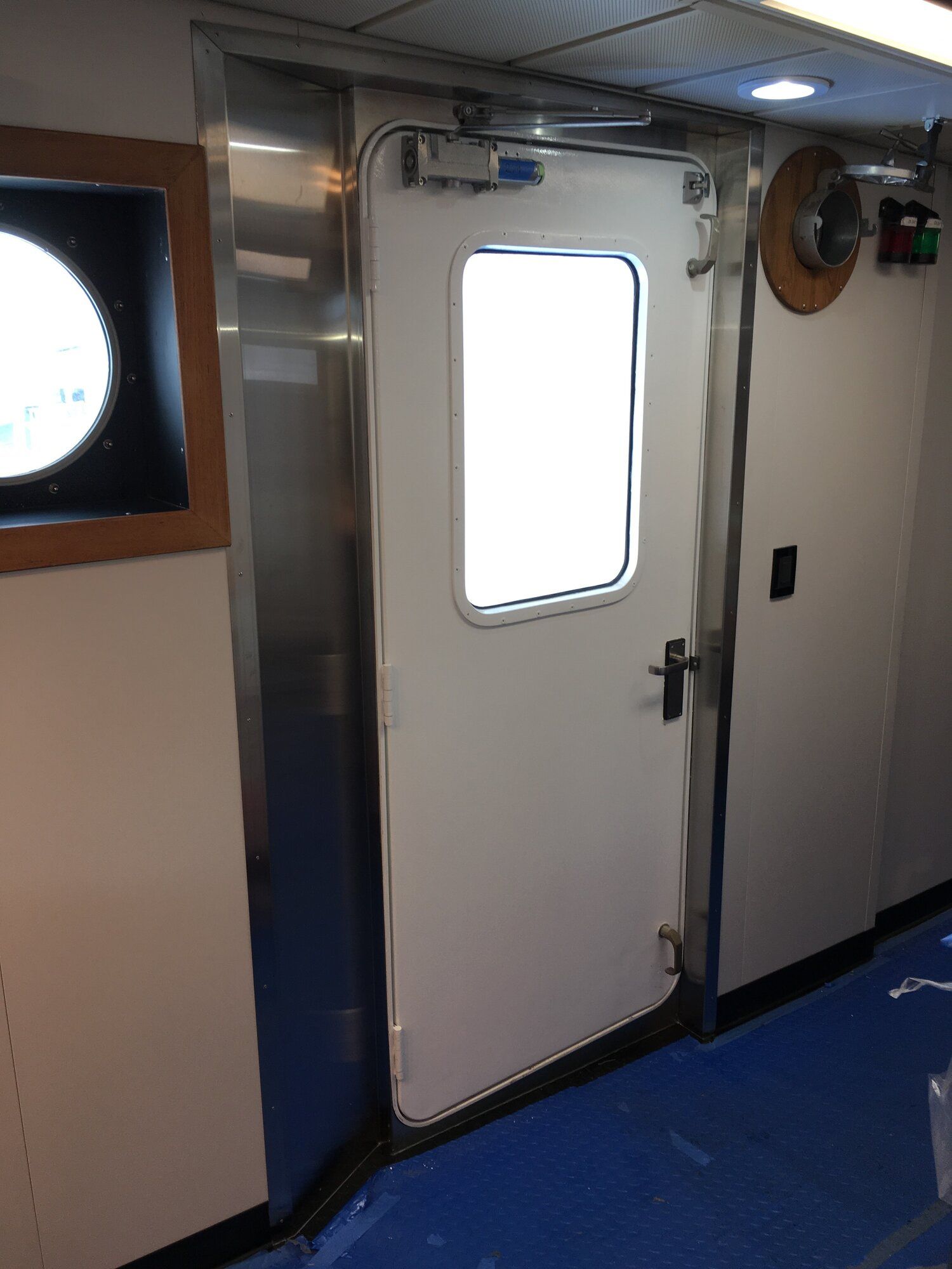 White door with window, on a boat, flanked by metal and white walls, blue floor.