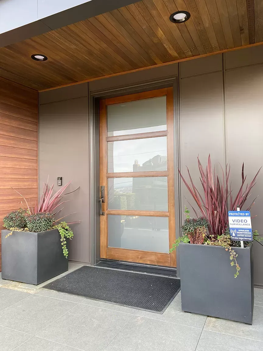 Modern home entrance with wooden door, glass panels, planters, and doormat.