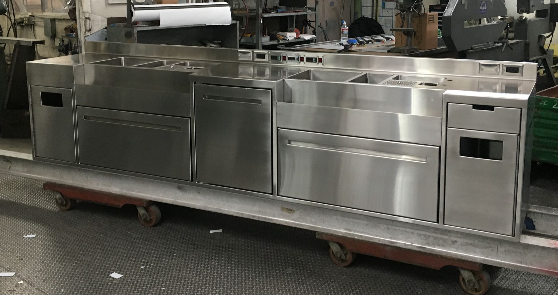 Stainless steel commercial kitchen counter with drawers and sinks on a rolling cart in a workshop.