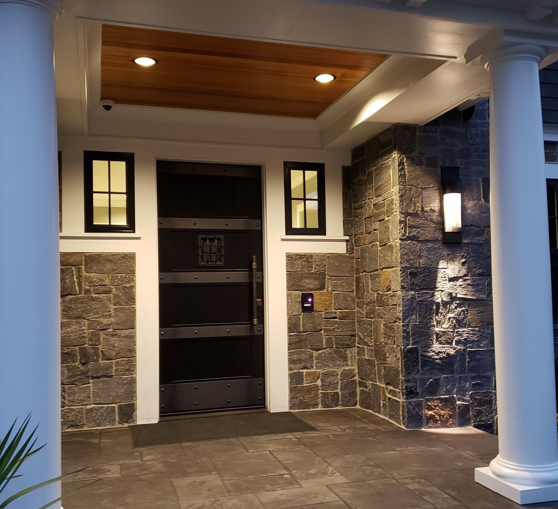 Entrance with a dark door, stone walls, and white pillars. Two small windows on each side of the door.
