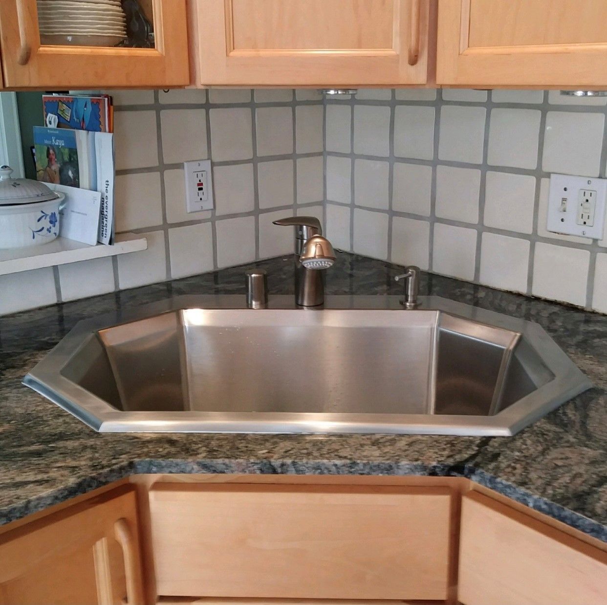 Stainless steel kitchen sink in a corner, surrounded by granite countertop and light-colored cabinets.