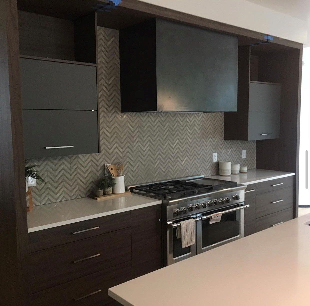 Modern kitchen with dark cabinetry, stainless steel oven, grey herringbone backsplash, and white countertops.