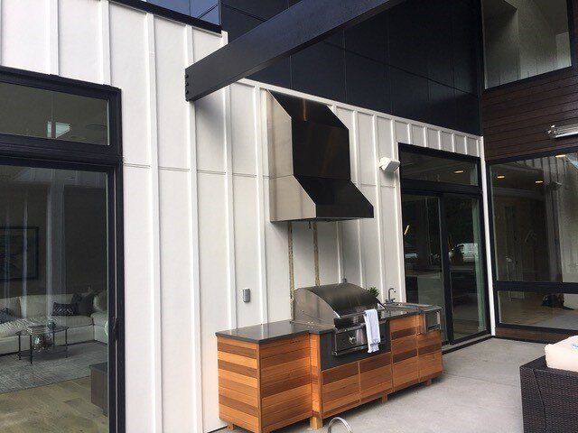 Outdoor kitchen with a stainless steel grill and a black hood. White siding, wood cabinets, and glass doors.