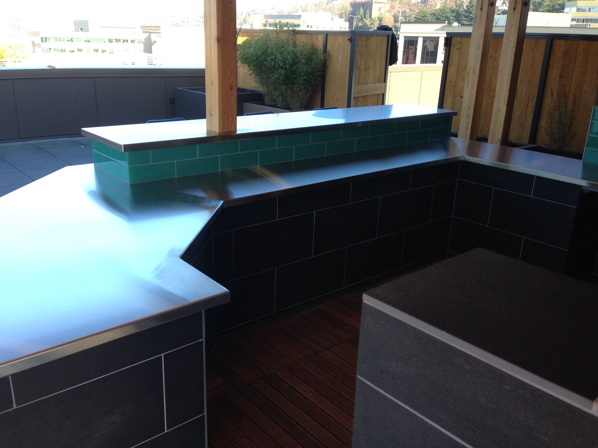Outdoor kitchen on a rooftop with stainless steel counters, dark tiled walls, and green tiled backsplash.