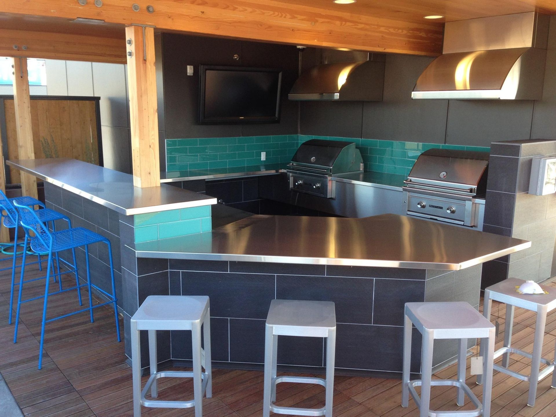 Outdoor kitchen with stainless steel countertops, grills, and teal tile backsplash. Includes seating.