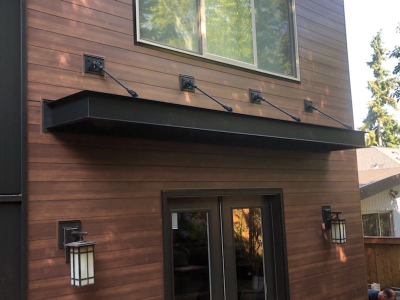 Black metal awning over a doorway, attached to a brown wooden house with wall lights.