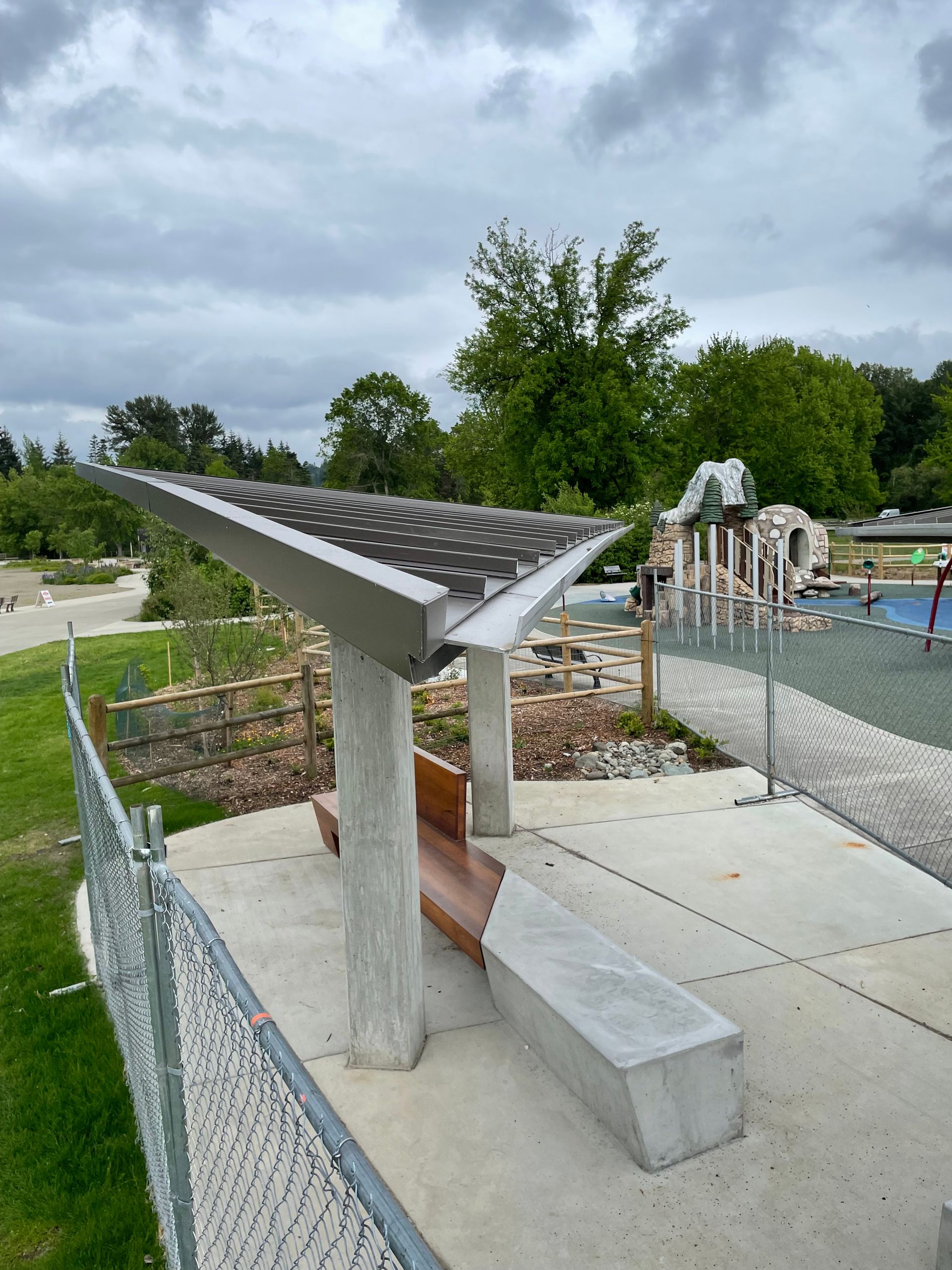 Park shelter with solar panel roof, concrete columns, wood bench, and playground in background.
