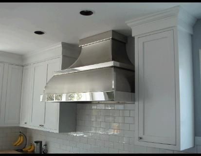 Stainless steel range hood over a white tiled backsplash and white cabinets in a kitchen.