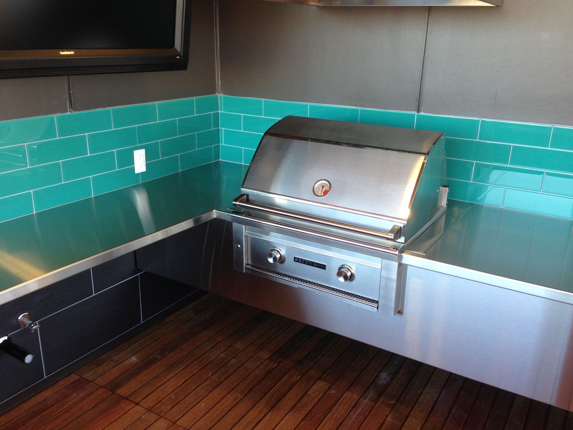 Stainless steel grill built into an outdoor kitchen with teal tile backsplash and wood flooring.