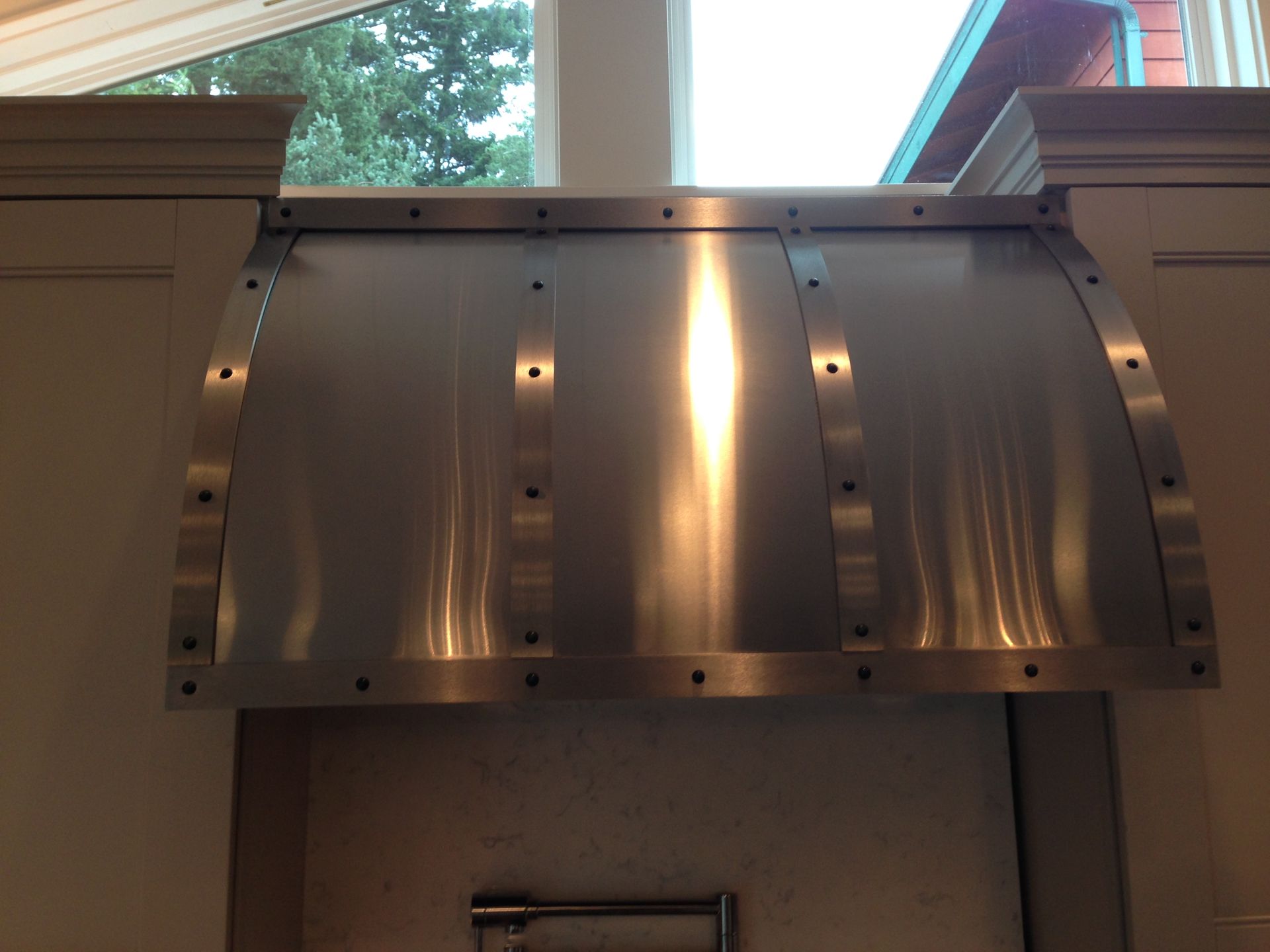 Stainless steel kitchen range hood with riveted straps, between cabinets.