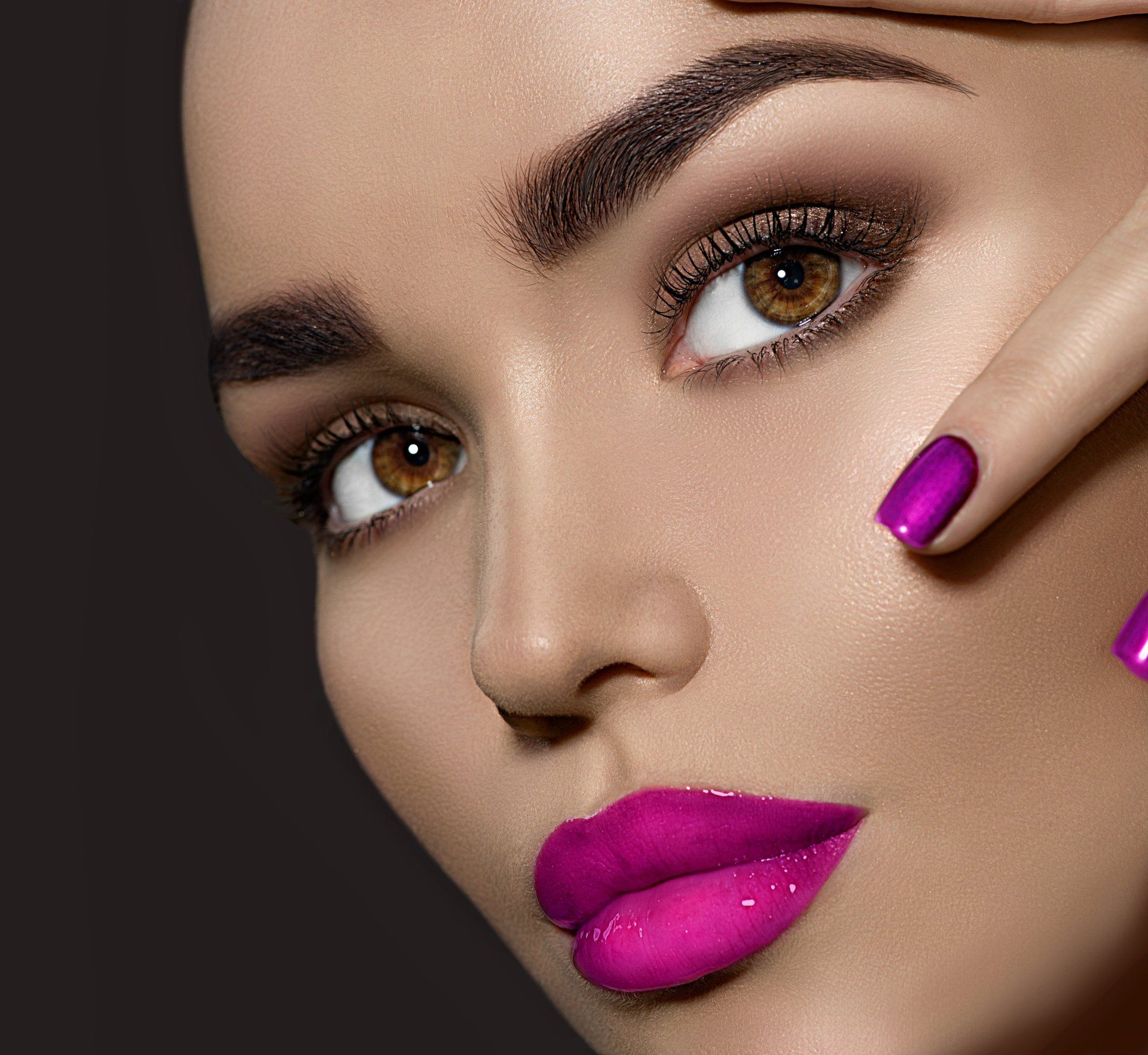 Woman with brown eyes and dramatic pink makeup; touching face with purple-nailed hand.