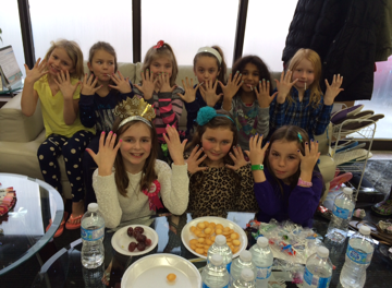 Ten young girls raise hands, showing off their painted nails, sitting on a couch in a room with snacks.