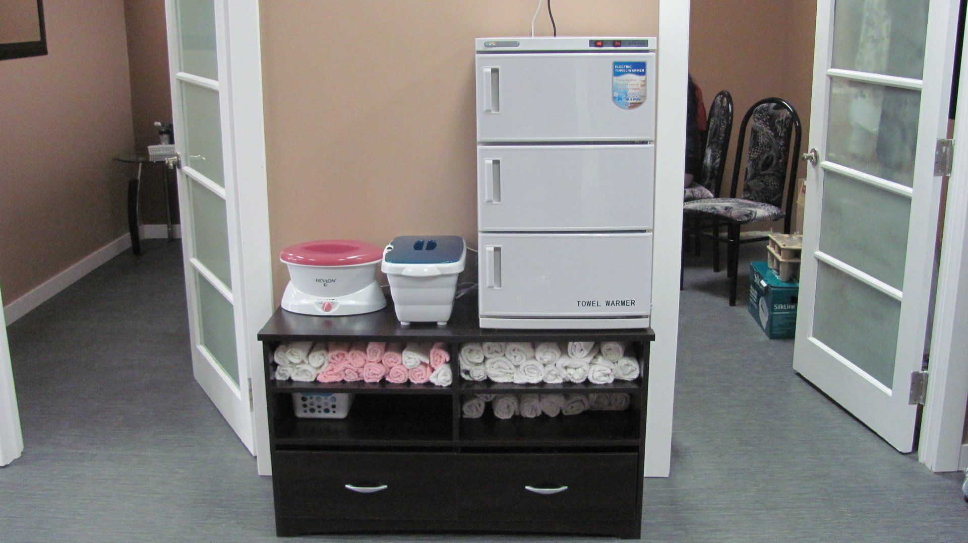 A spa towel warmer, wax warmer, and towels sit on a dark cabinet between two doorways.