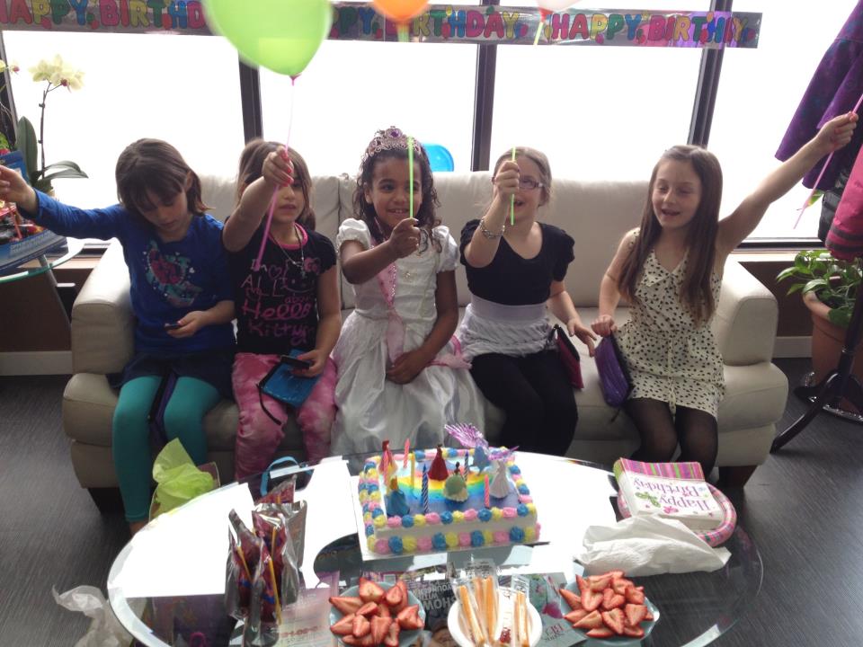 Five girls holding wands over a birthday cake, sitting on a couch. Balloons and colorful decorations.