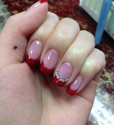 Hand with red-tipped French manicure, one nail adorned with silver gems, on a red and brown speckled surface.