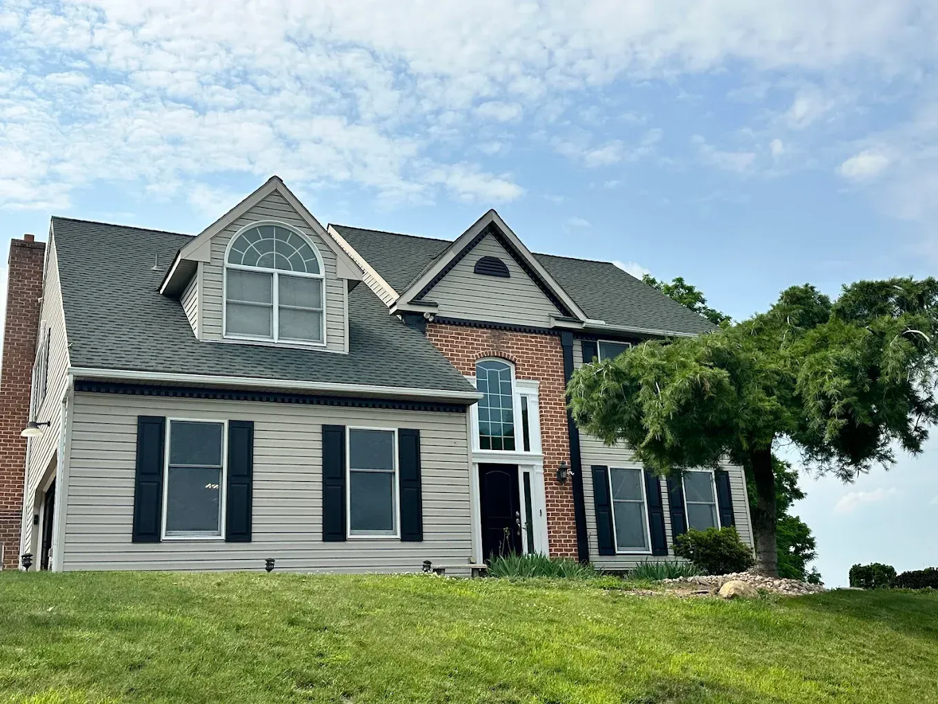 Two-story house with light gray siding, dark shutters, and a green lawn under a blue sky.