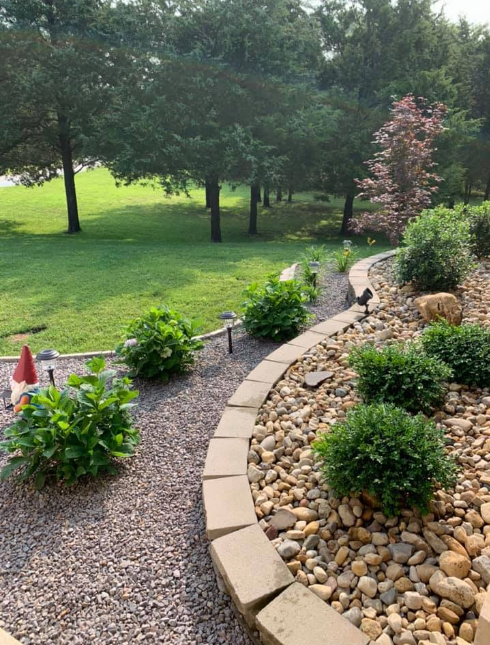 A garden with rocks , bushes and trees in the background created by Hackmann Lawn & Landscape STL