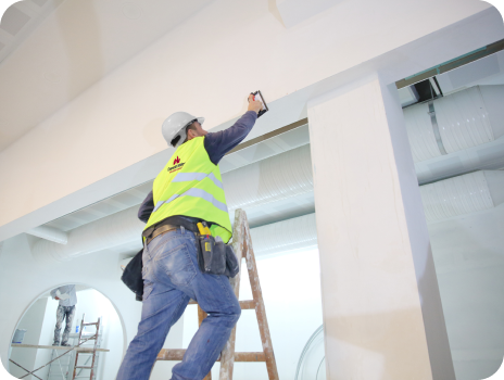 A man is standing on a ladder measuring a wall.