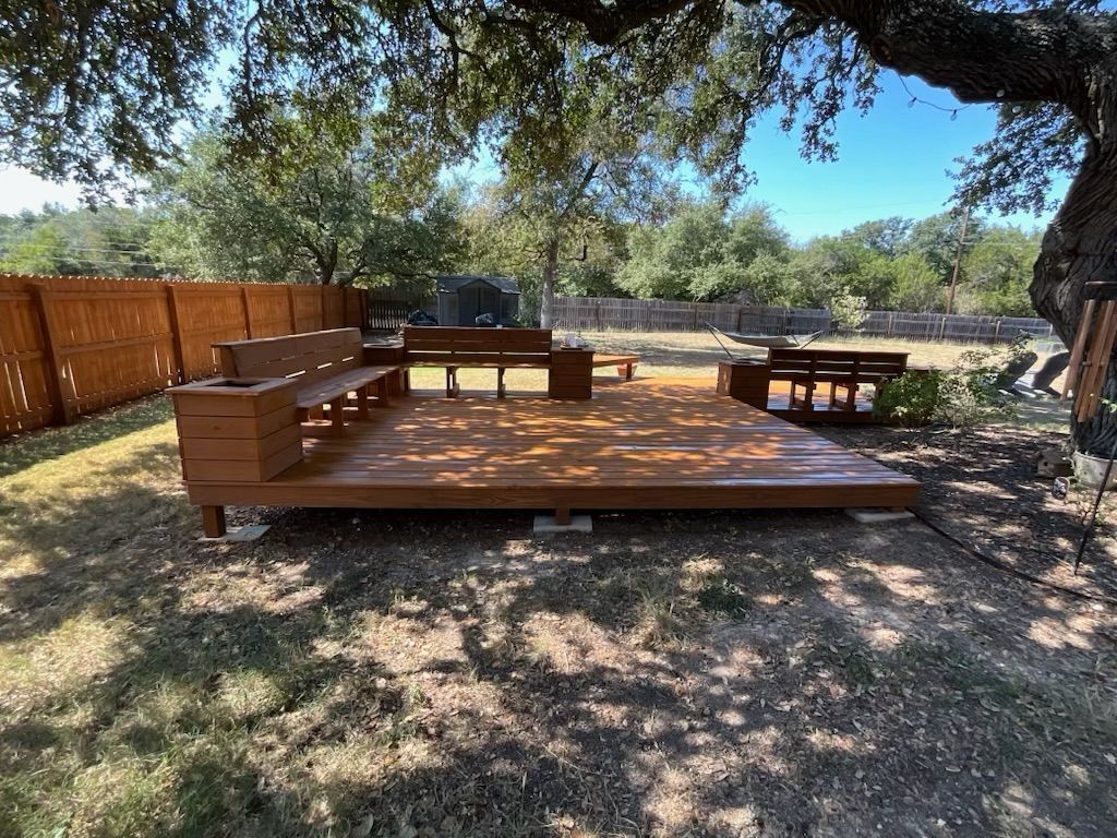 A wooden deck with a bench under a tree in a backyard.