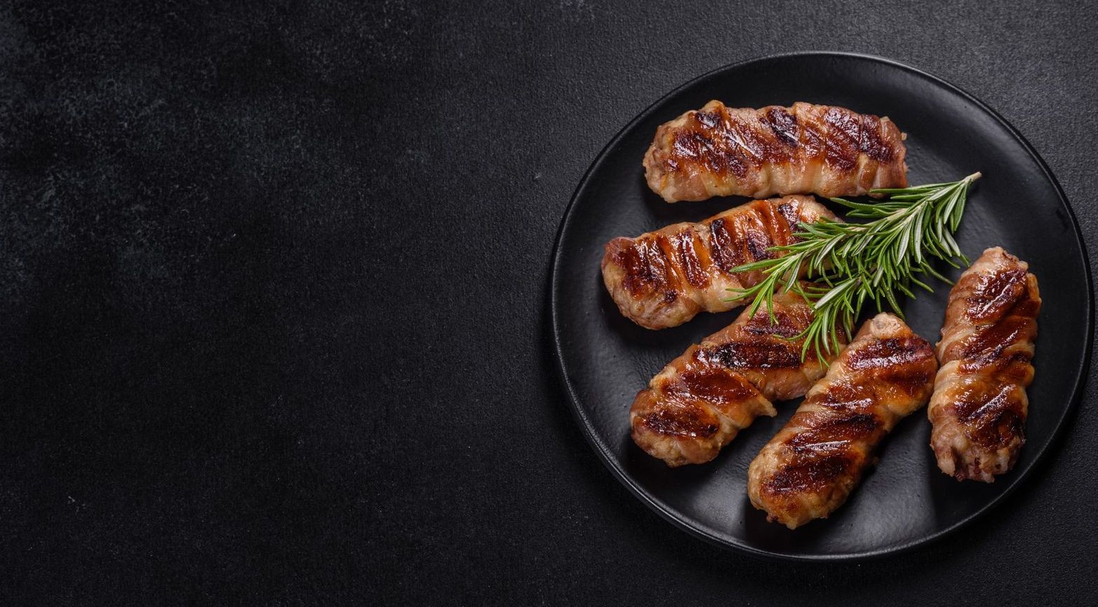 Salsicce grigliate su un piatto nero con rosmarino su sfondo nero.
