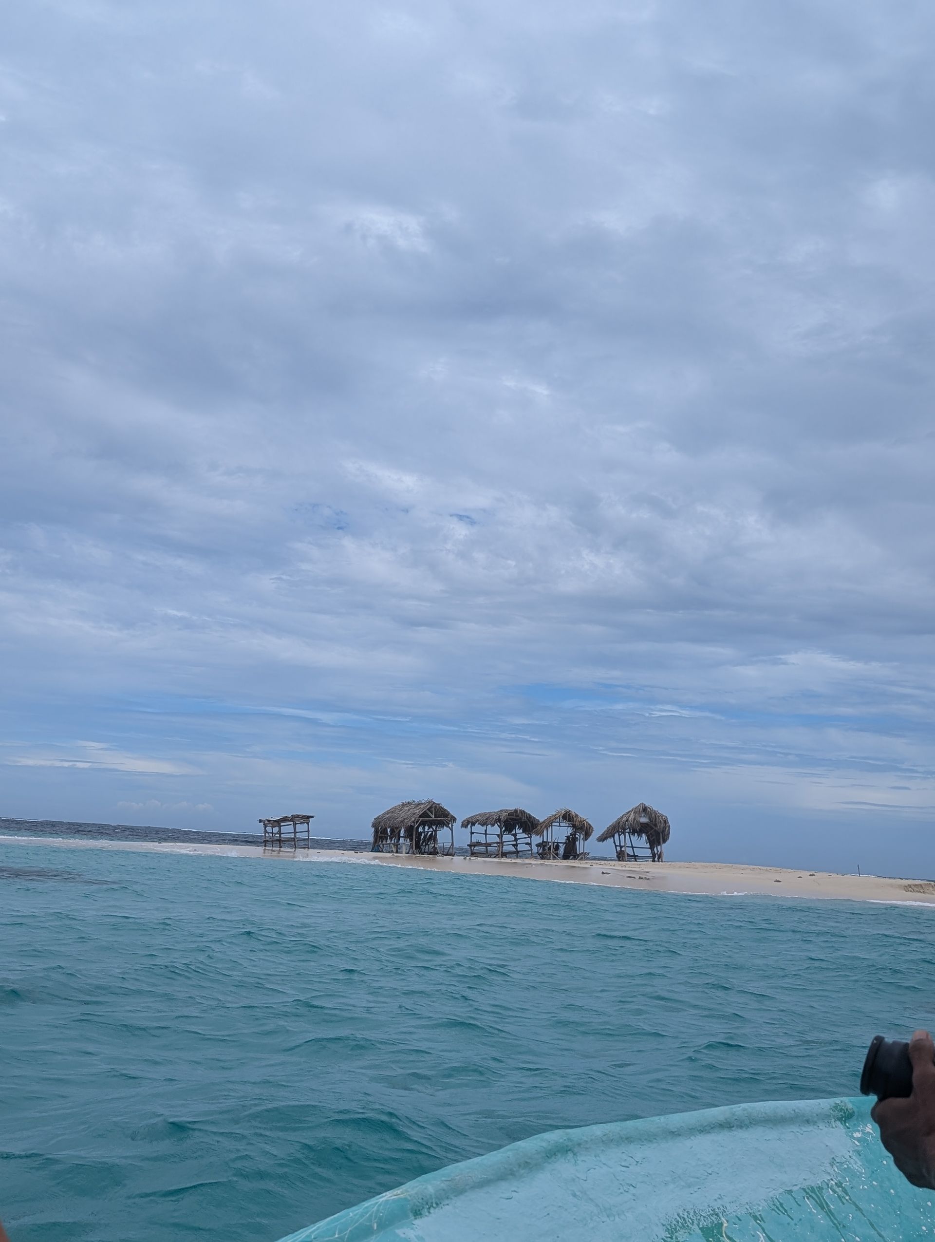 View from a boat of small huts on a sandbar in the ocean under a cloudy sky.