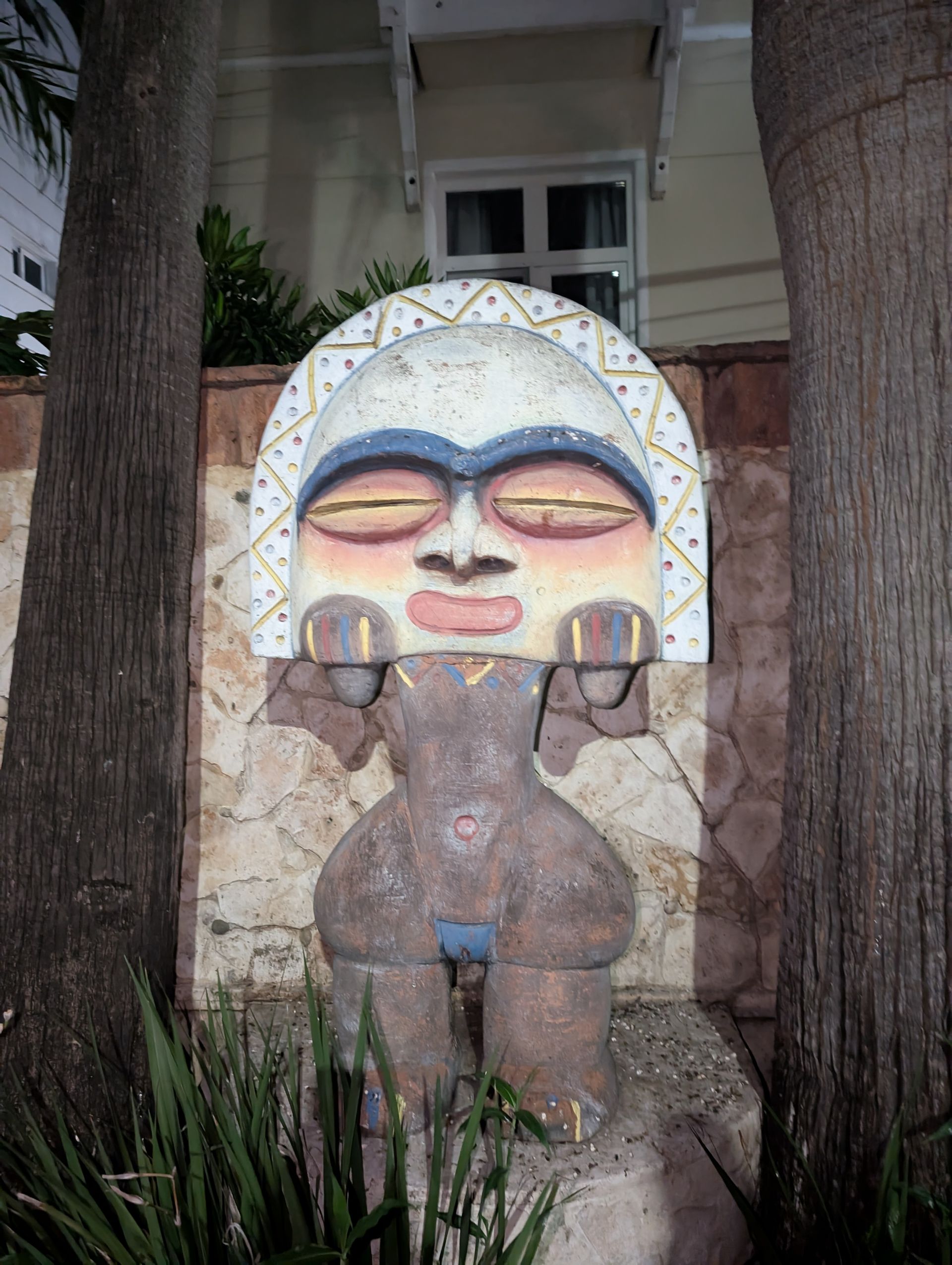 Stone sculpture of a stylized figure with large head and stylized features, mounted on a textured wall, flanked by trees.