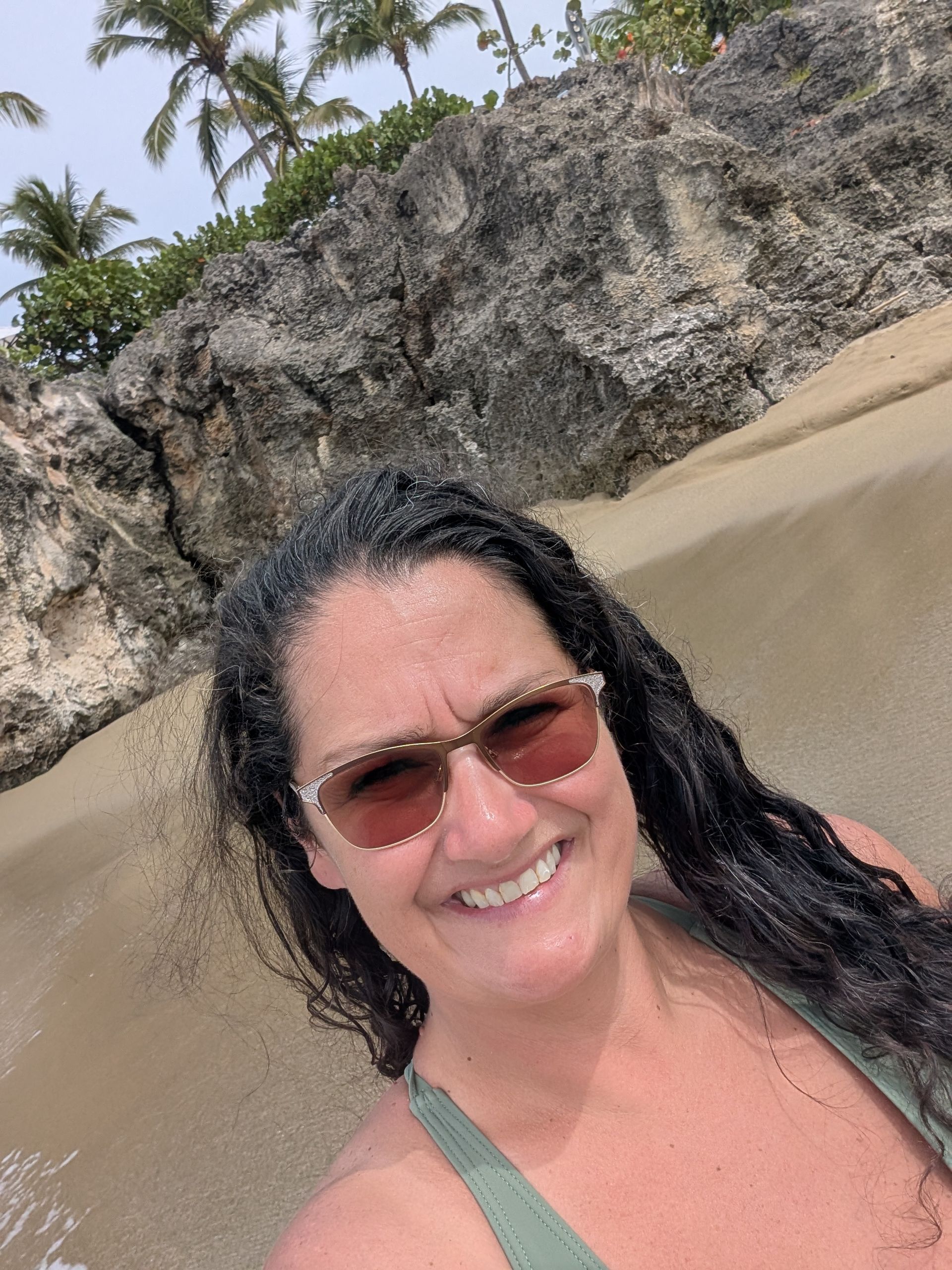 Woman in sunglasses smiles on a sandy beach near rocky cliffs, palm trees in the background.