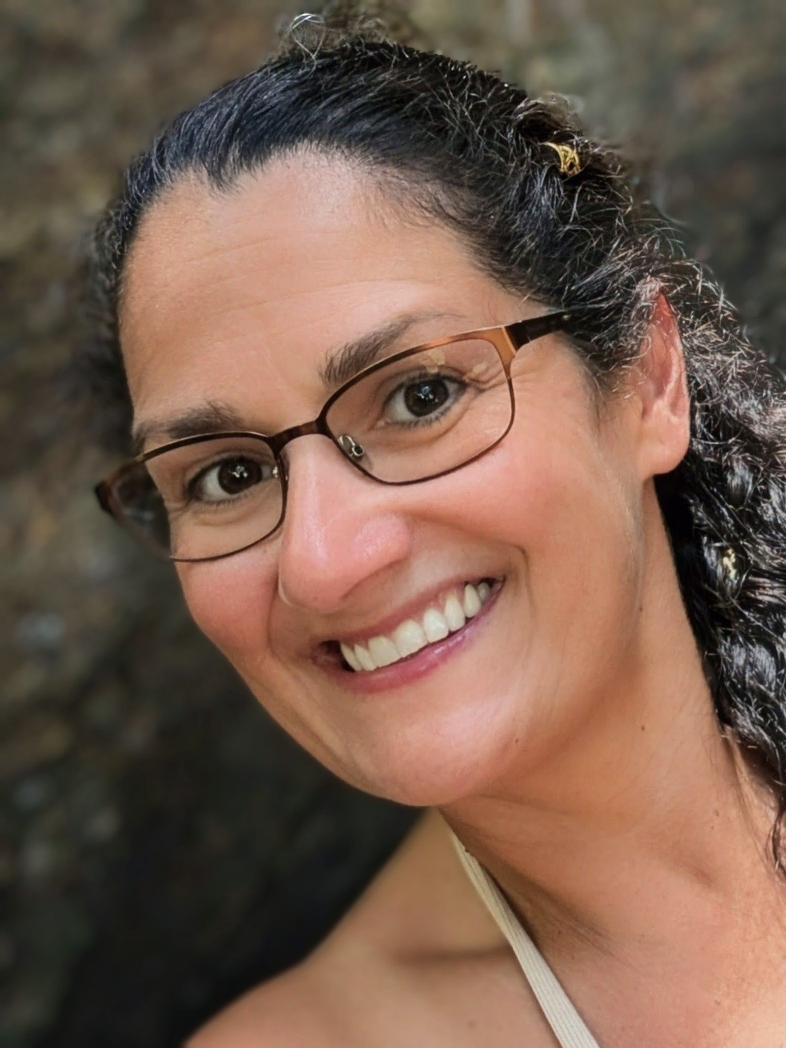 Woman smiling, wearing glasses and a white strap, outdoor background.