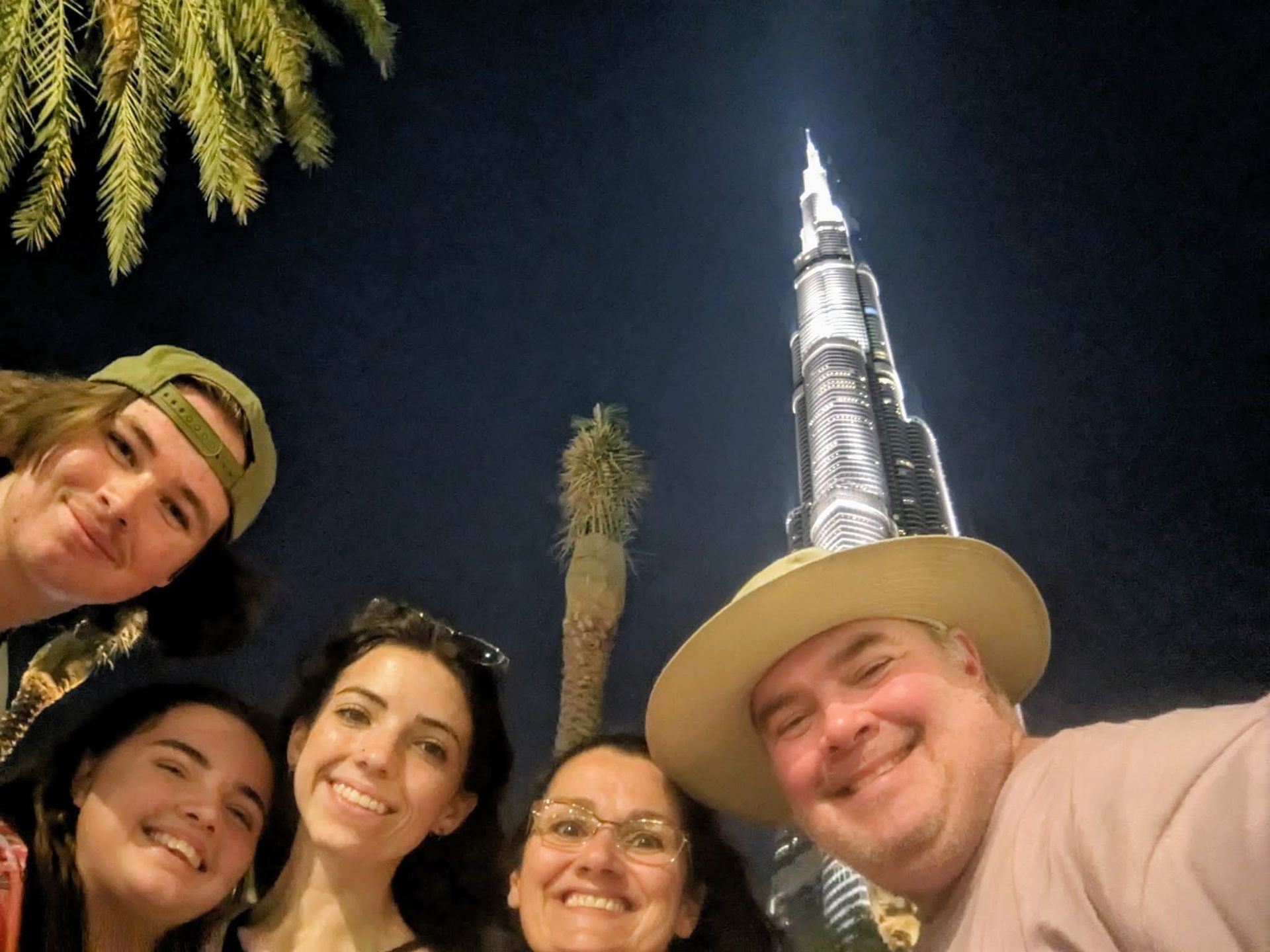 Family selfie in front of the Burj Khalifa at night. Palm trees frame the skyscraper, people smiling.