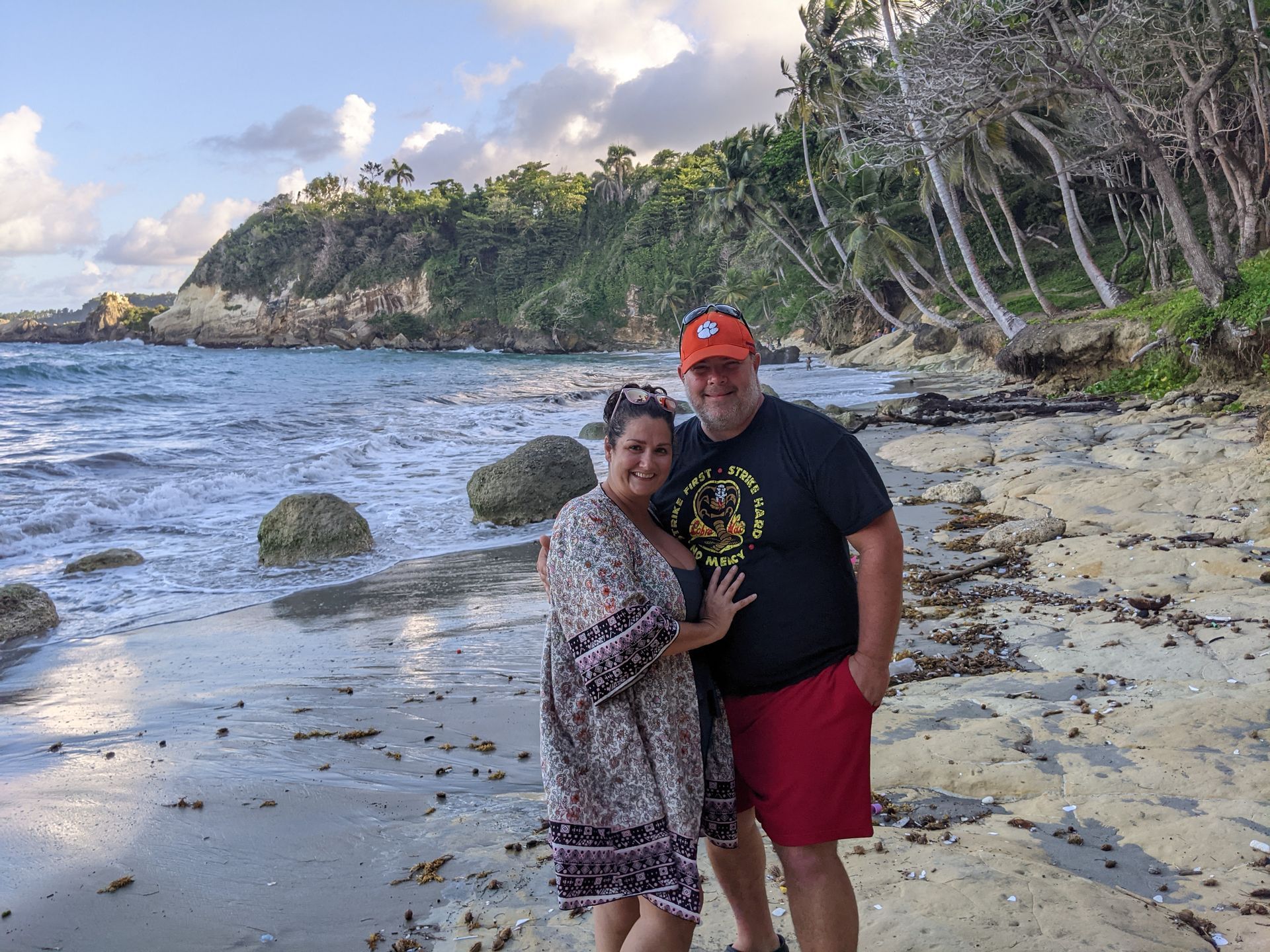 Couple on a beach, near water and green cliffside, man in red shorts and black shirt, woman in dress.