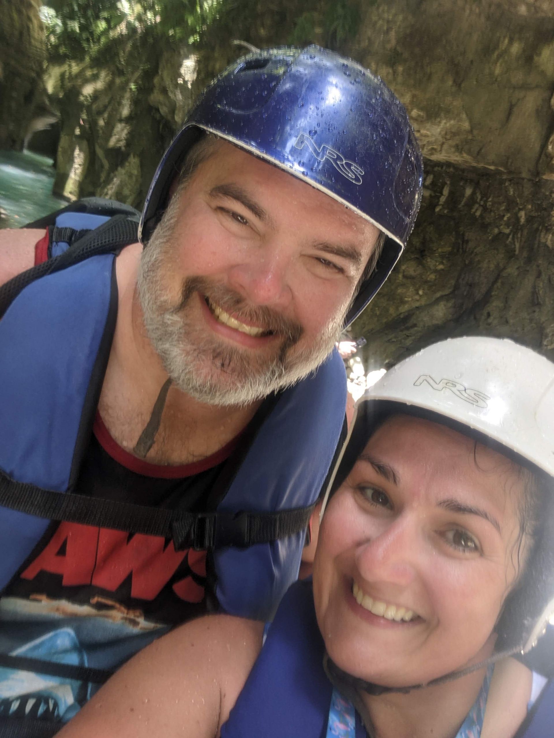 Couple in helmets and life vests smile, possibly canyoning or rafting. Blue, rocky background.