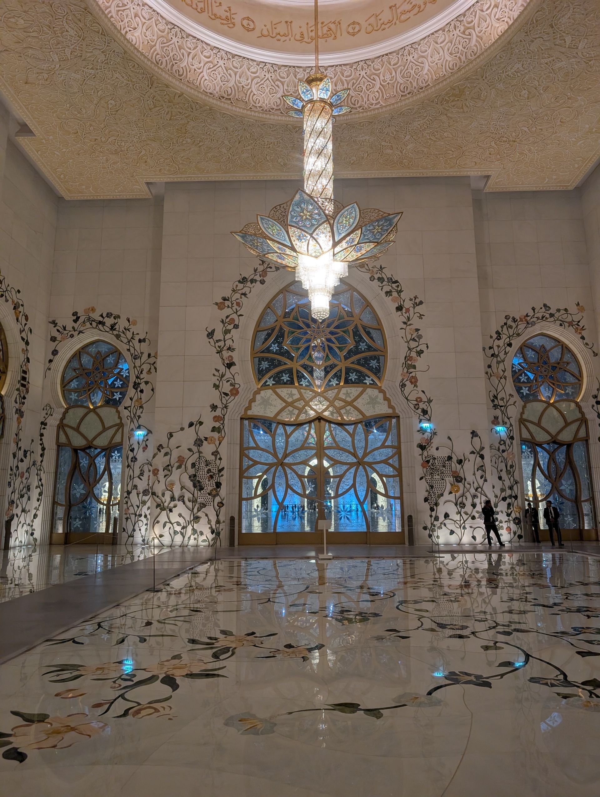 Interior of mosque with ornate floral design, large chandelier, and tiled floor.