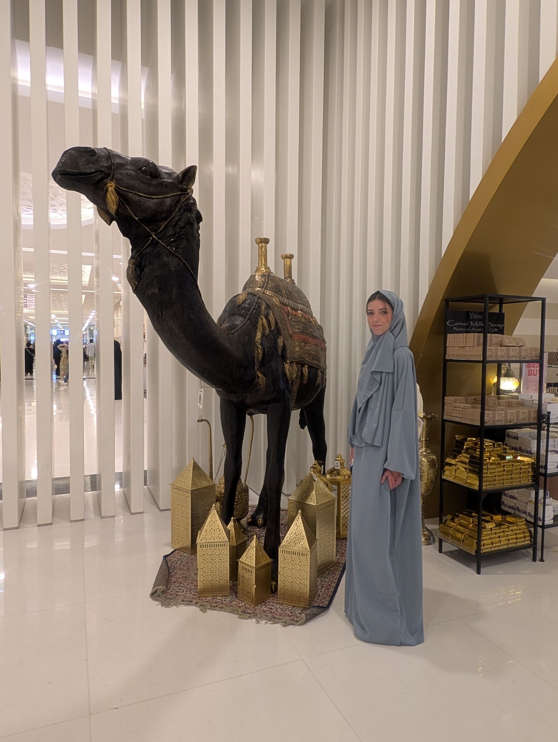 Person in light blue garment stands beside a dark camel sculpture in a bright interior.