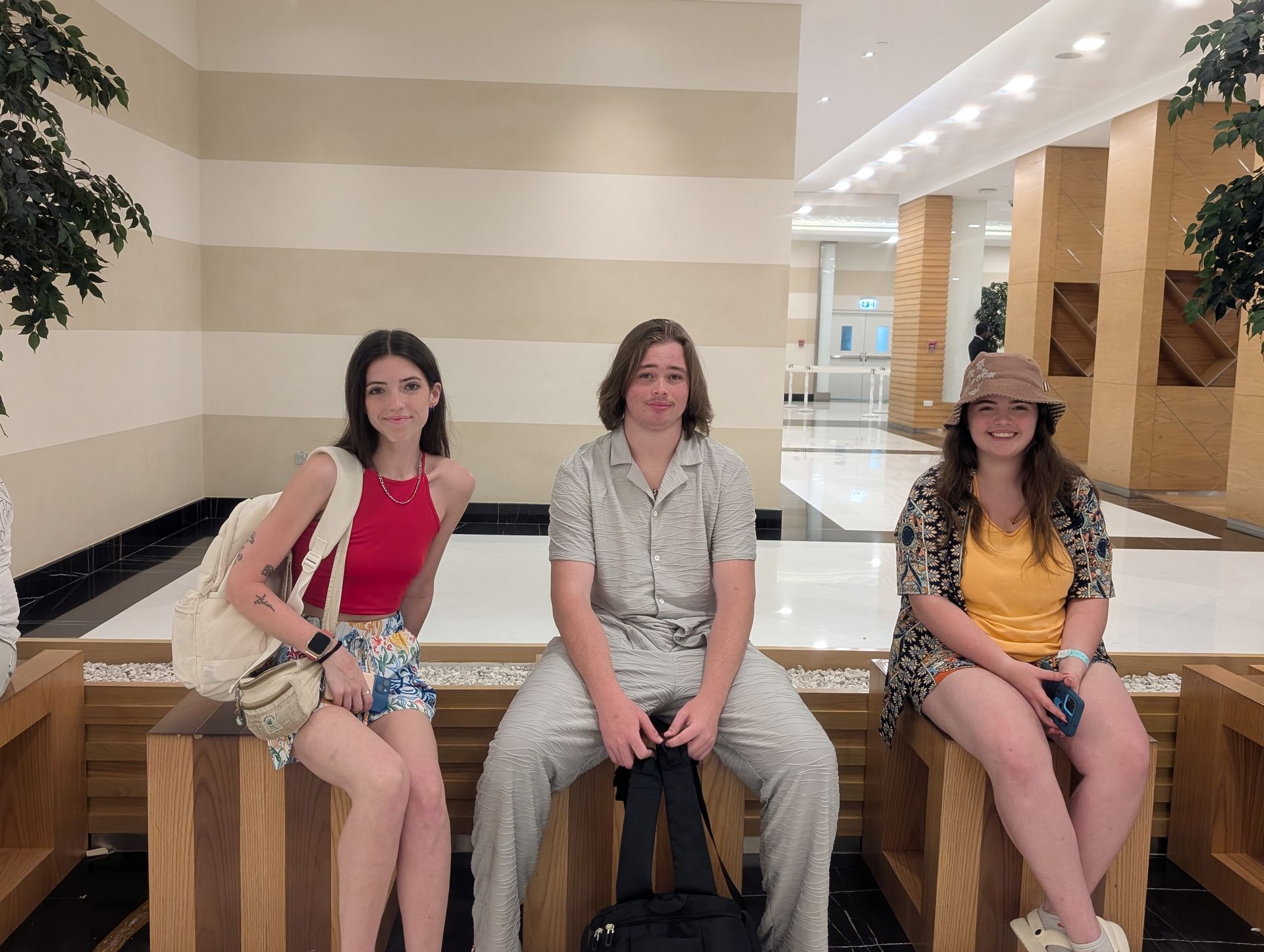 Three people sitting on a wooden bench indoors. The center person holds a bag; the others have bags and phones.