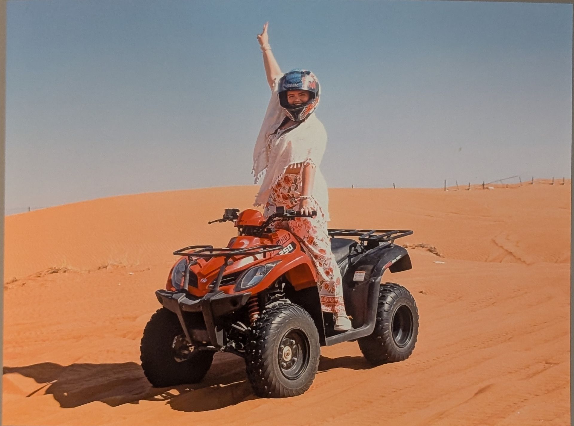 Person on an ATV, waving in a desert setting. The vehicle is red, against tan sand and a blue sky.