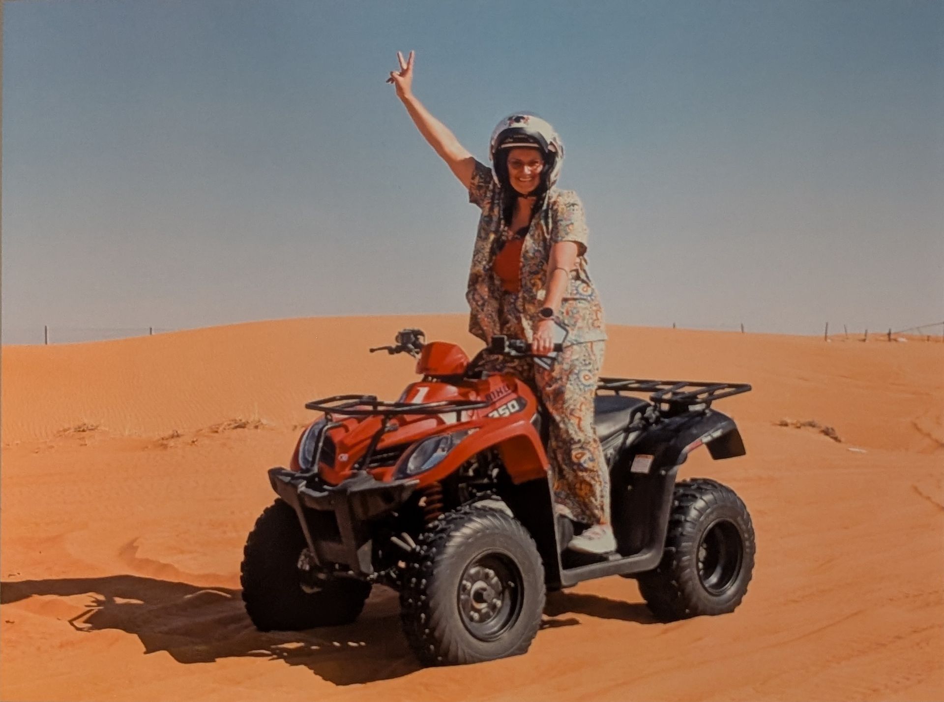 Person on ATV in desert, raising hand. Red vehicle, tan sand, blue sky.