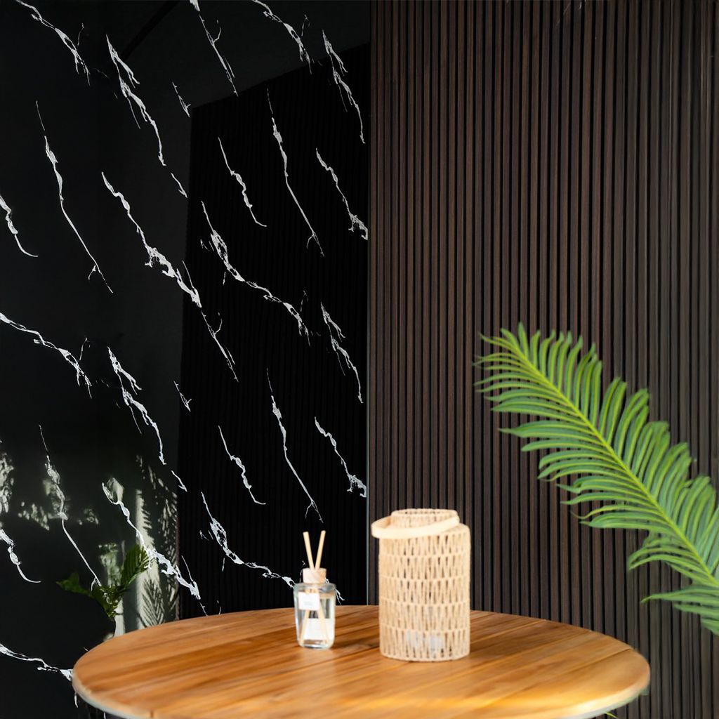 Mesa minimalista con farol tejido, difusor de caña y hoja de palma verde sobre una pared negra con paneles de madera.
