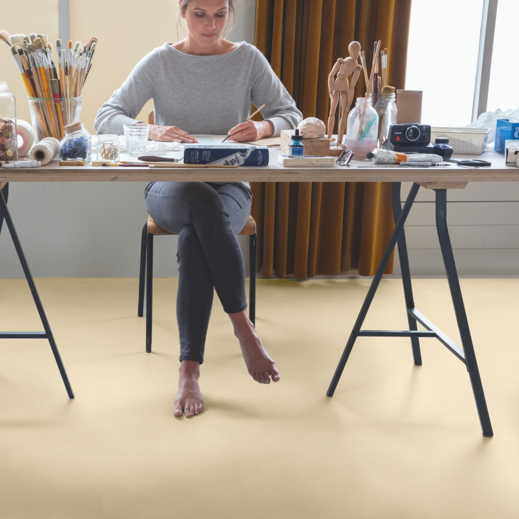 Una artista dibuja en una mesa desordenada en un estudio luminoso, rodeada de pinceles, figuritas y materiales de arte.