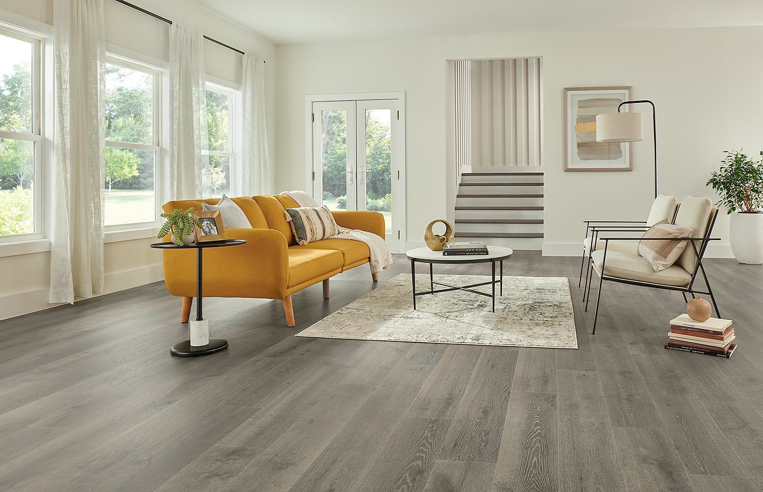 Luminoso salón con suelo de madera gris, sofá color mostaza, silla blanca, alfombra, mesa de centro y grandes ventanales.