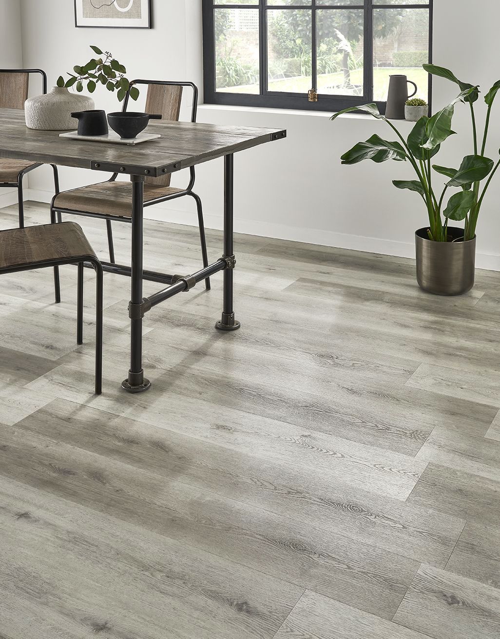 Comedor moderno con mesa rústica, sillas negras, plantas de interior y suelo de madera clara junto a una ventana.