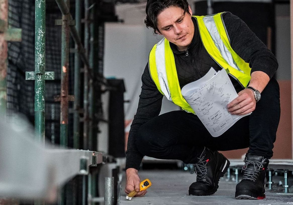 A construction worker is squatting down while holding a tape measure.