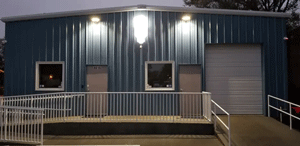 Blue metal building with two doors, windows, and overhead lights; a ramp and railing lead to the entrance.