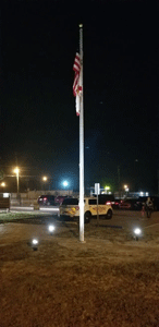 American flag at half-staff on pole at night; lights illuminate lawn and vehicles.