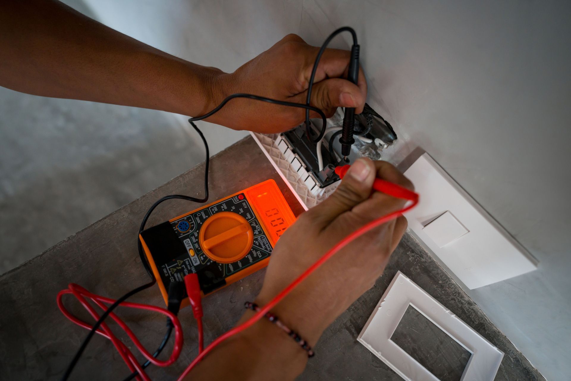 Hands using a multimeter to test a wall outlet, orange meter, white walls.