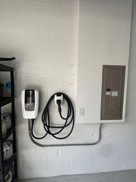 White wall-mounted EV charger and electrical panel in a garage. Black cable runs in a metal conduit.