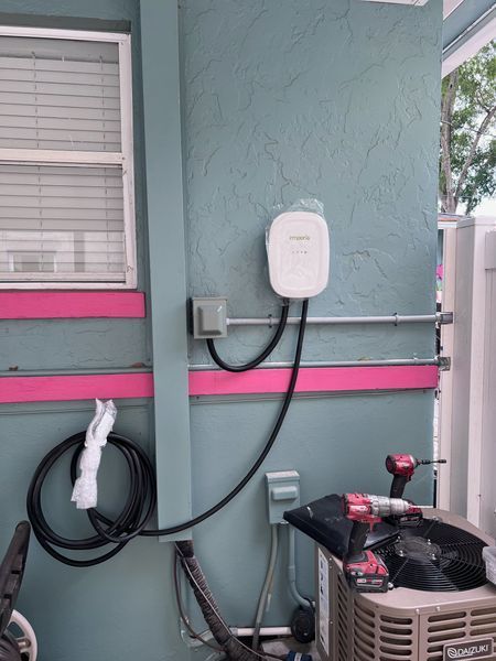 Electric vehicle charger mounted on an exterior blue wall with pink trim. Electrical conduit and tools are visible.