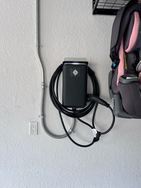 Black EV charger mounted on white wall next to electrical conduit and a car seat.
