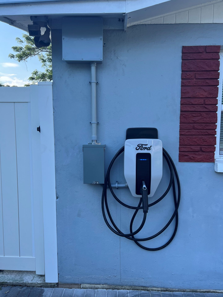 Ford electric vehicle charger mounted on a light blue wall with electrical conduit and a red shutter.