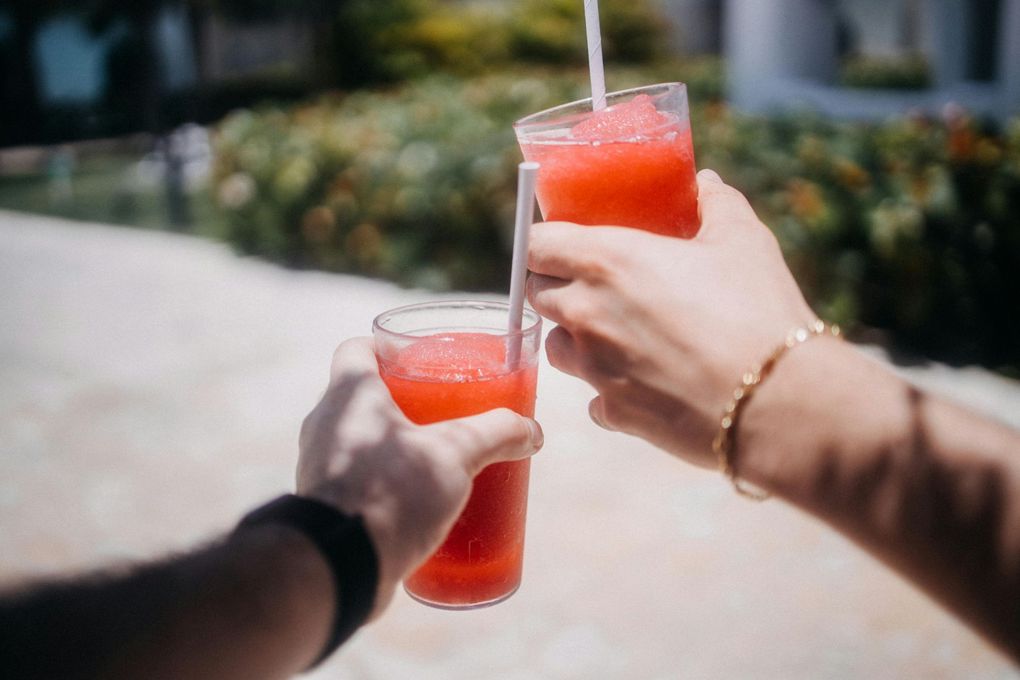 Two people clink glasses filled with red frozen drinks, outdoors on a sunny day.