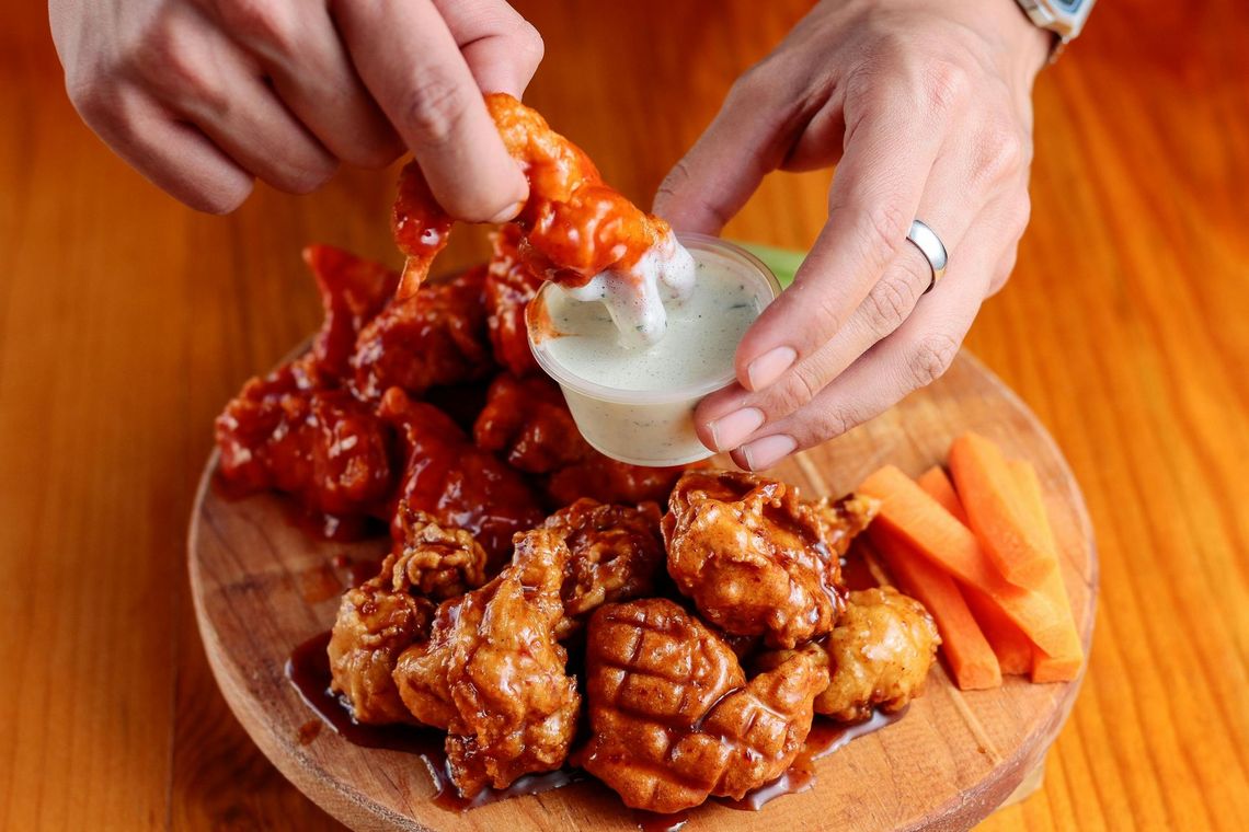 Person dipping a buffalo wing into ranch dressing on a wooden plate with other wings and carrot sticks.